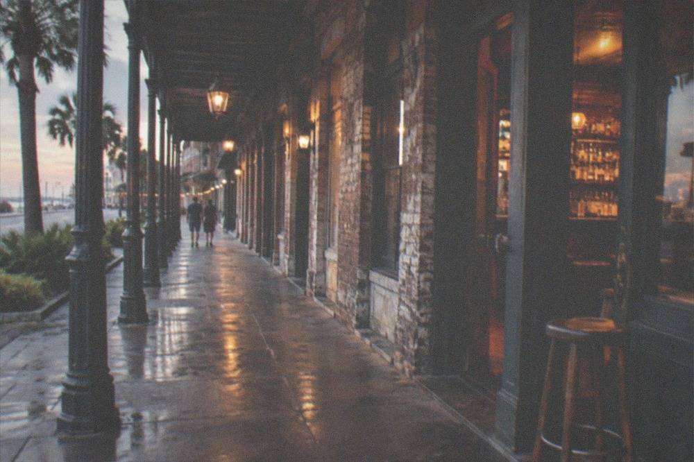 A Victorian era commercial street of brick storefronts lit by evening lamps with a bar sign visible