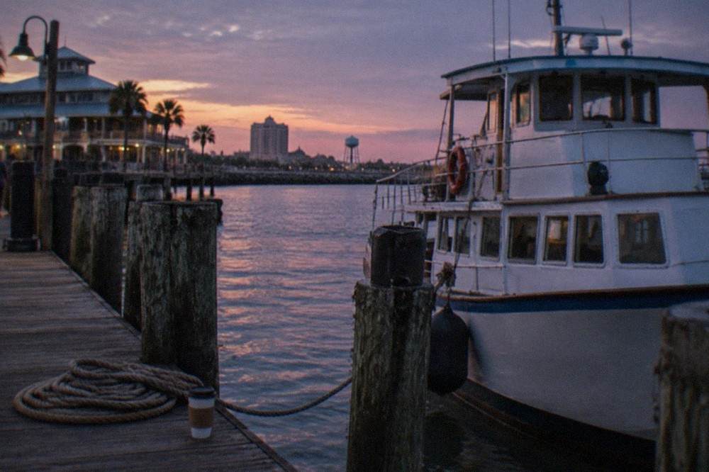 A tour boat at a Galveston pier at dusk with dark harbor water and docked vessels behind it