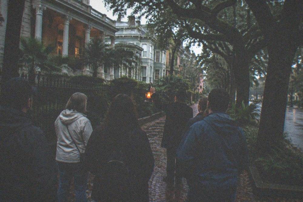 A small group of people standing on a sidewalk listening to a tour guide at night